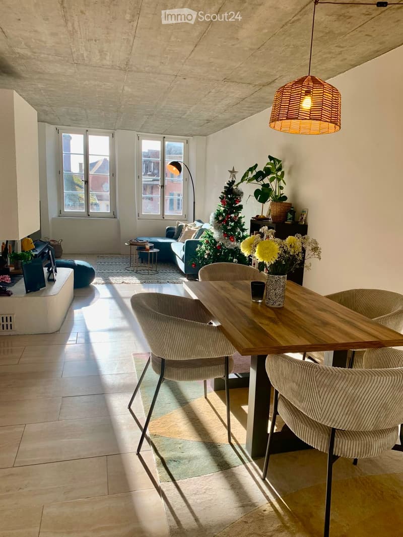 Bright living room with large windows, a sofa, a wooden dining table with chairs, a pendant light, a floor lamp, a Christmas tree, and decorative vases.