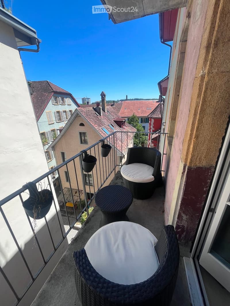 Balcony with a round table and two wicker chairs, overlooking a cityscape with traditional houses and a clear blue sky.