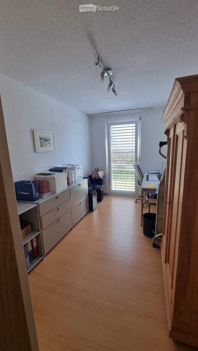 Office space with wooden flooring, large window with blinds, desk with chair, filing cabinet, and various office supplies.