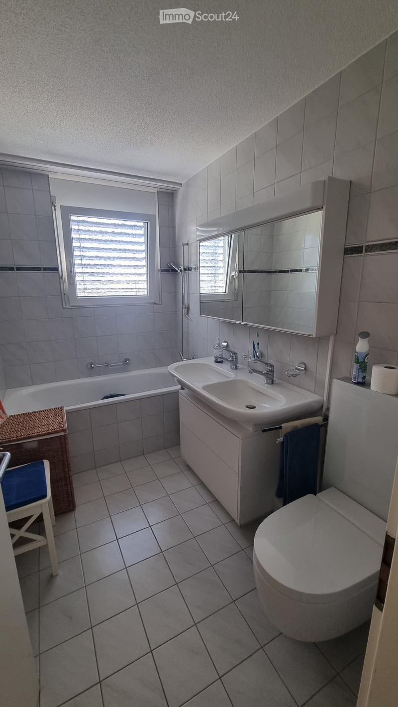 Modern bathroom with a bathtub, double sink, toilet, mirror, and a small chair near the window.