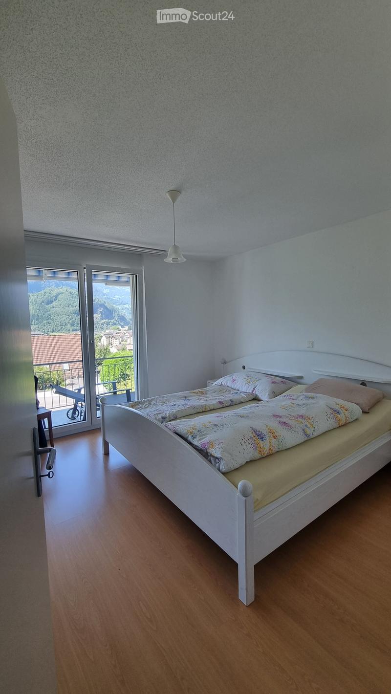 Bright bedroom with a white bed, colorful bedding, and a nightstand, featuring large windows with a scenic mountain view.