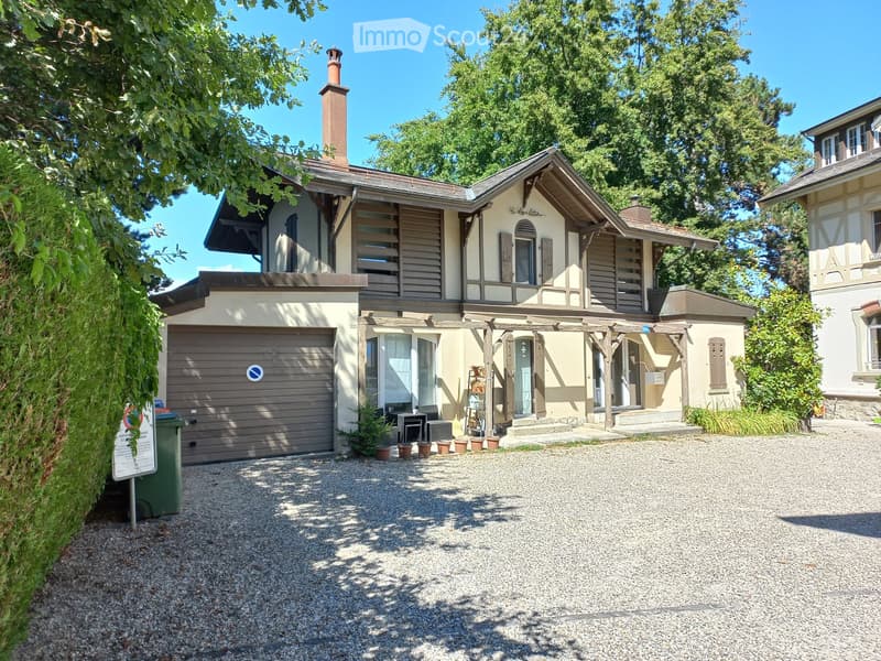 Two-story house with a garage, surrounded by greenery and a gravel driveway.