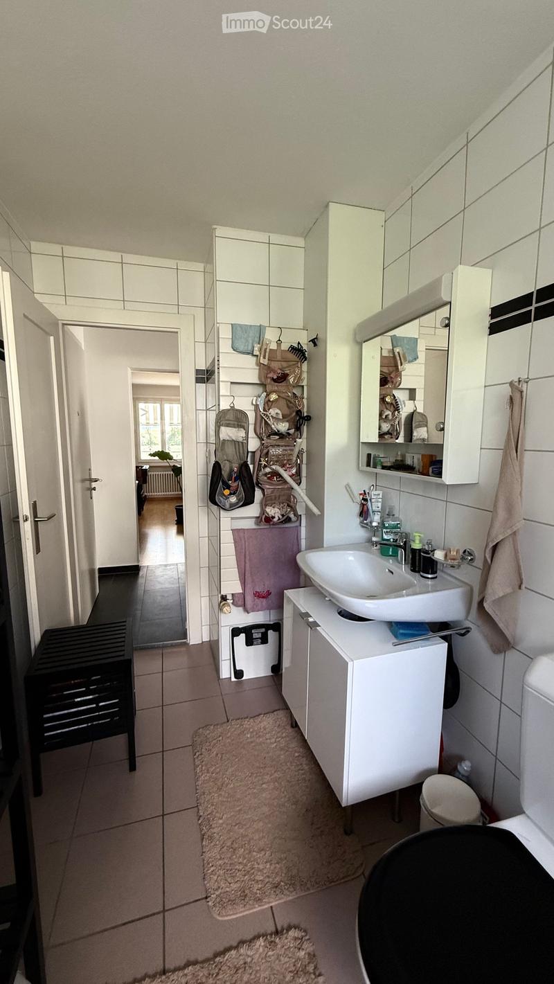 Modern bathroom with white tiles, a sink, mirror, cabinet, toilet, and towels. A small stool is visible near the door.
