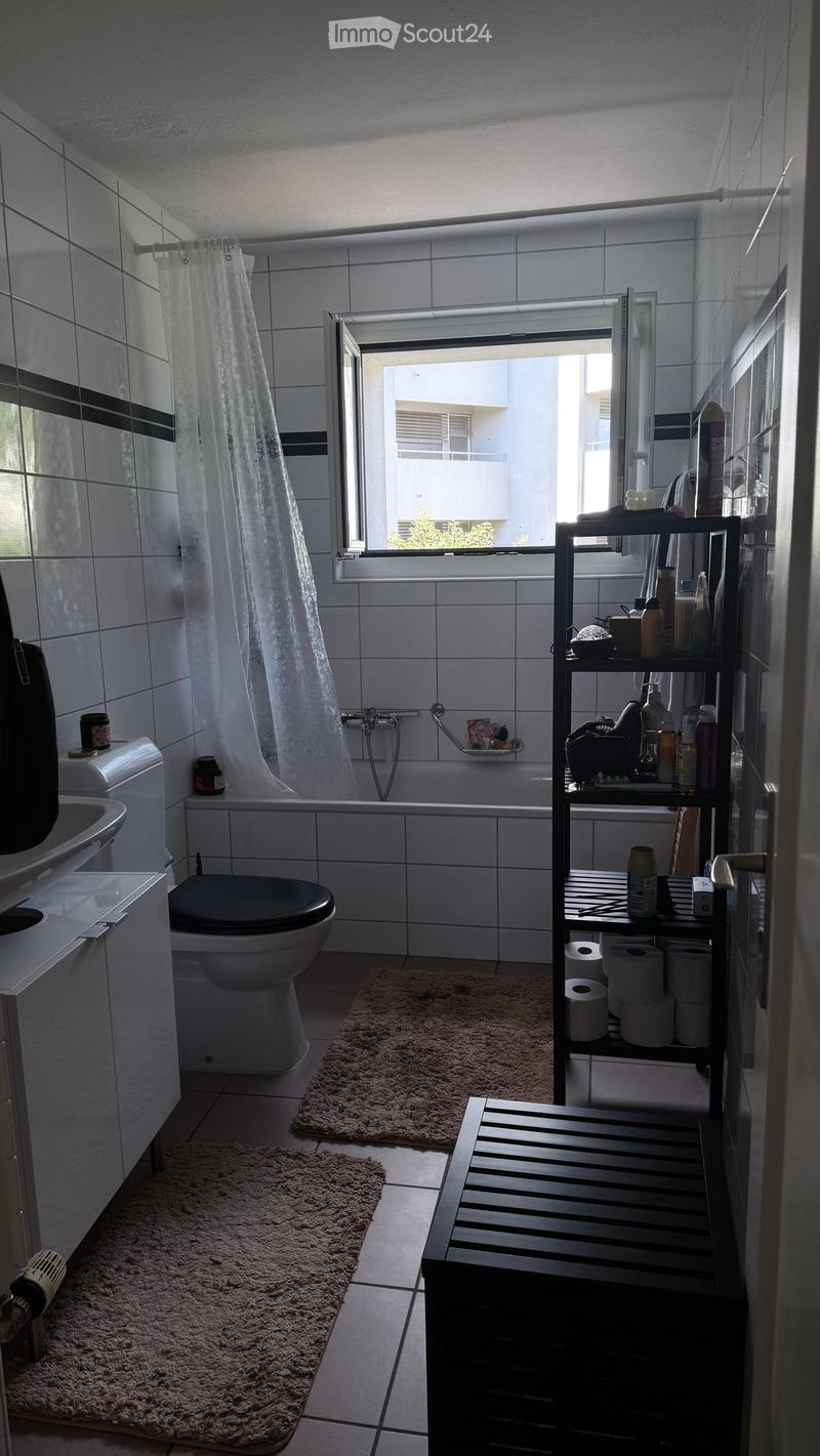 Modern bathroom with white tiles, a toilet, bathtub, sink, shower curtain, shelf with toiletries, and two rugs on the floor.