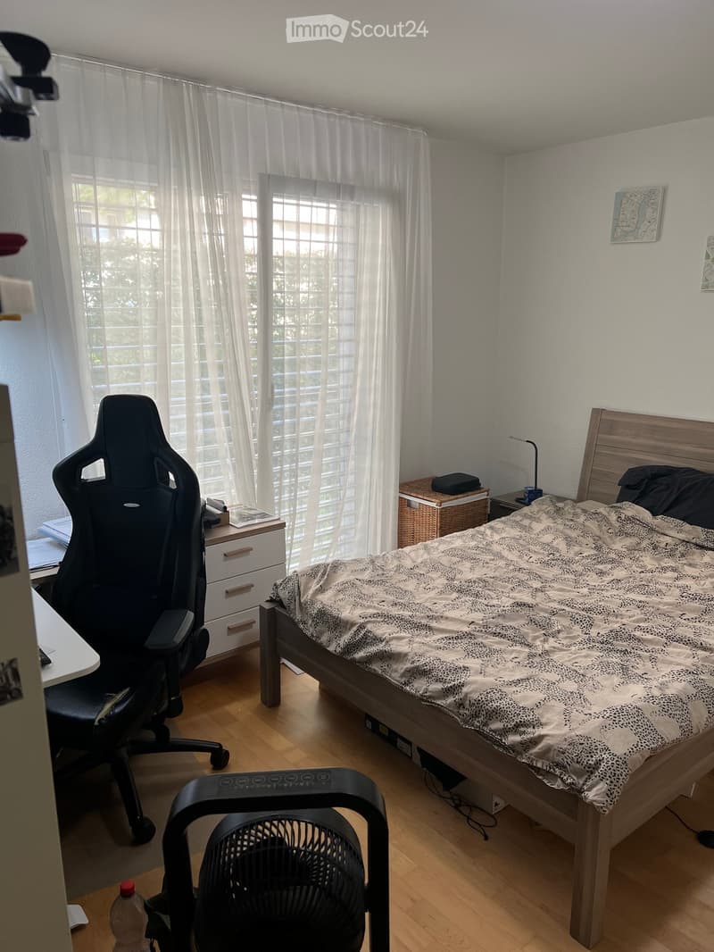 A well-lit bedroom with a wooden bed, a patterned duvet, a black gaming chair, a small table with a lamp, and a fan on the floor.