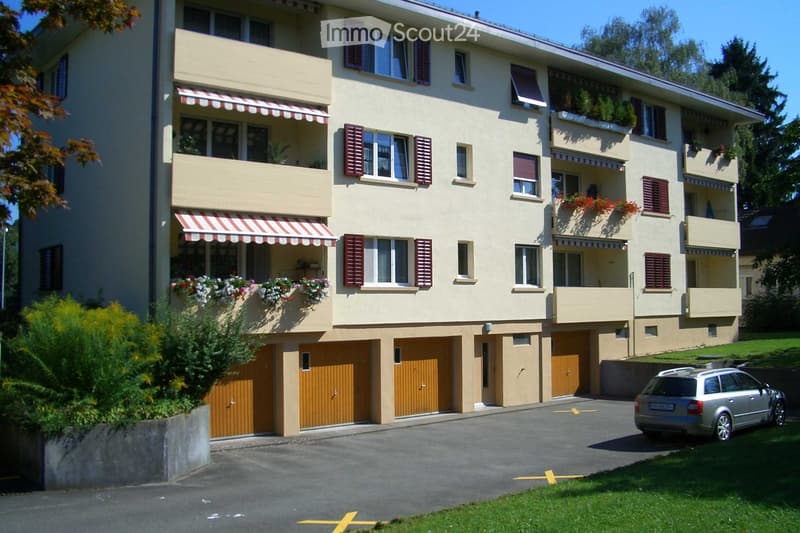 A multi-story residential building with beige walls, red shutters, balconies with flower boxes, and a parking area with a car.