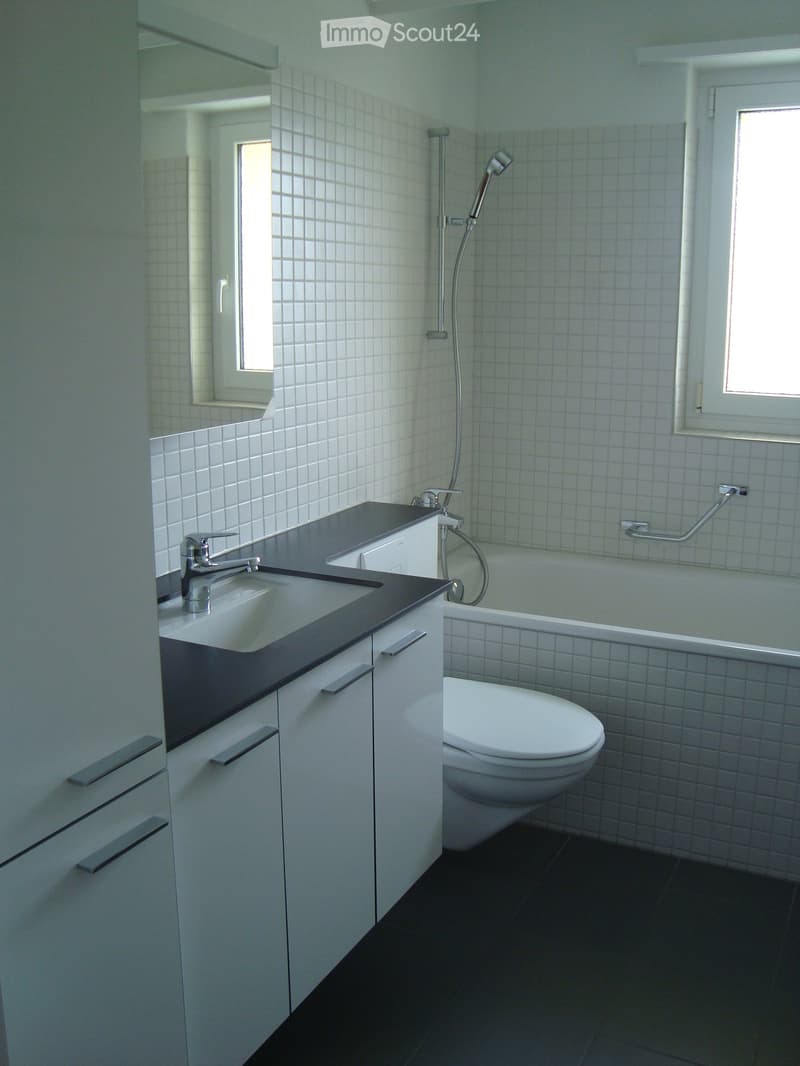 Modern bathroom with white tiled walls, dark countertop, sink, toilet, bathtub, and showerhead.