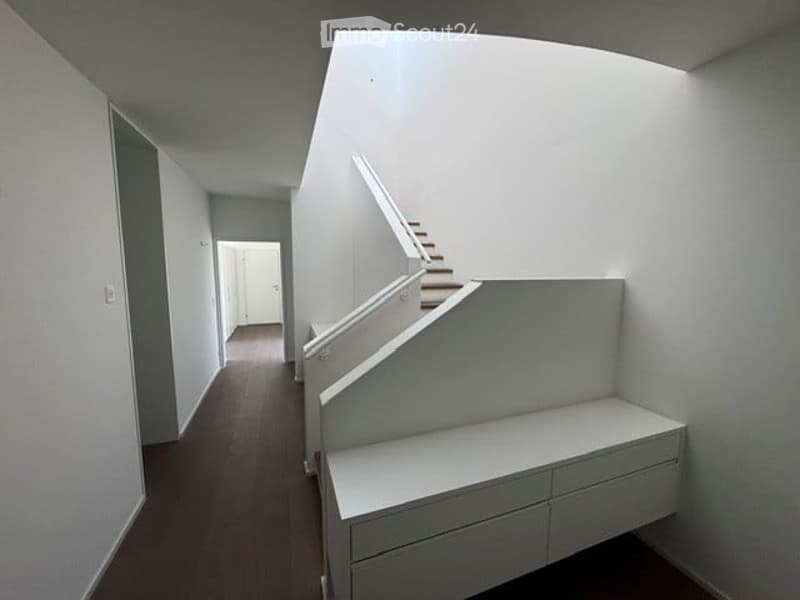 Modern hallway with white walls, wooden flooring, and a staircase leading upwards. The hallway features built-in storage units and a neutral color palette.