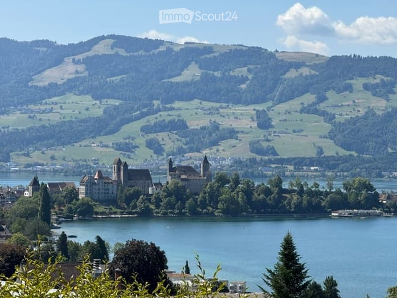 A historic castle sits on an island in a lake, surrounded by lush green hills and a clear blue sky.