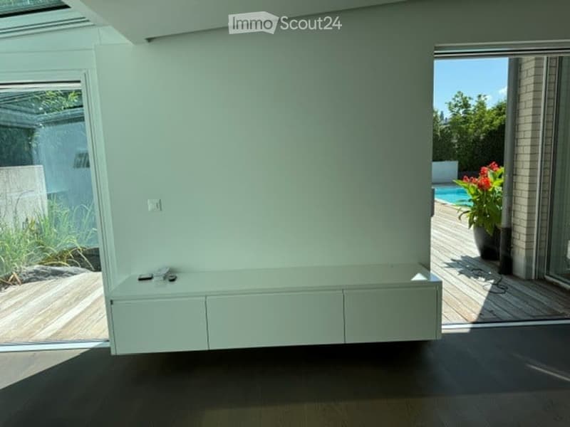 Bright, minimalist room with white walls, dark flooring, and built-in white cabinetry. Large windows and sliding glass doors lead to an outdoor deck with a swimming pool and greenery visible.