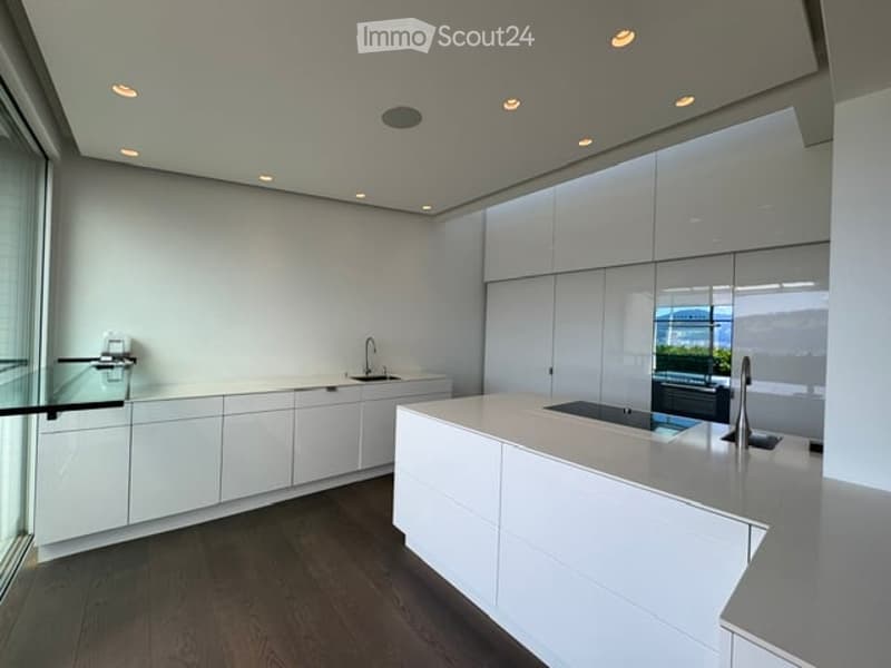 Modern, minimalist kitchen with white cabinets, a central island, and a sleek countertop. The space features recessed lighting and large windows offering a view of the outdoors.