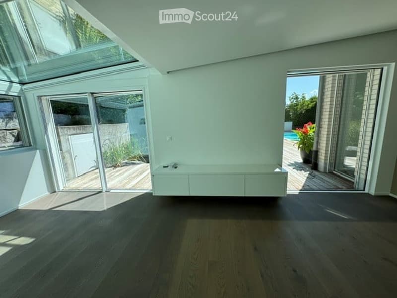 Bright, empty room with large glass sliding doors leading to a wooden deck and outdoor area, featuring a built-in white bench along one wall and a dark wooden floor.