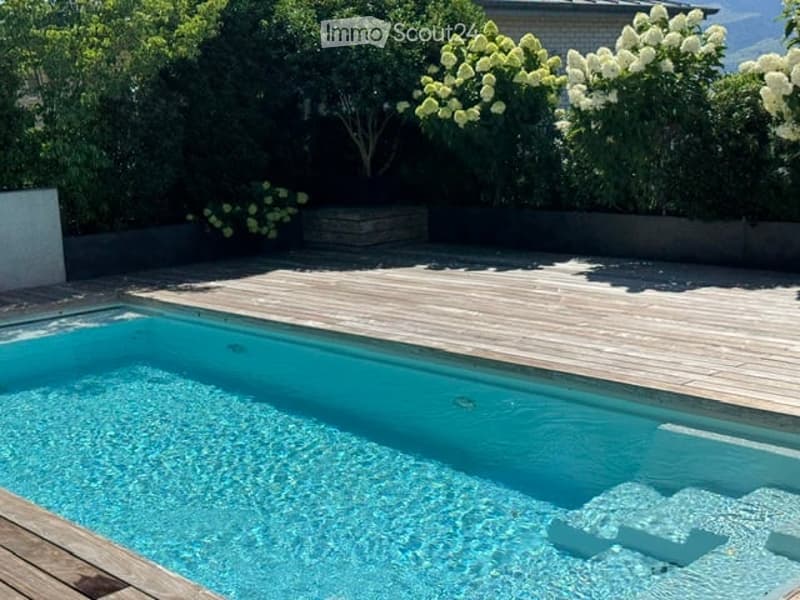 A modern outdoor area featuring a rectangular swimming pool with clear blue water, surrounded by a wooden deck and lush greenery, including flowering shrubs and trees.