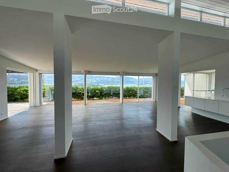 Open-plan unfurnished room with large windows and a view of the landscape, featuring a wooden floor and white walls.