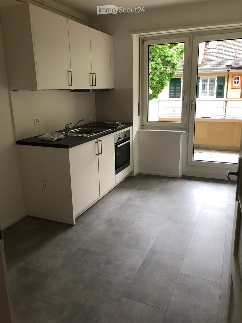 Modern kitchen with white cabinets, black countertop, sink, and oven. Large sliding glass door leads to a balcony with a view of greenery and neighboring buildings.