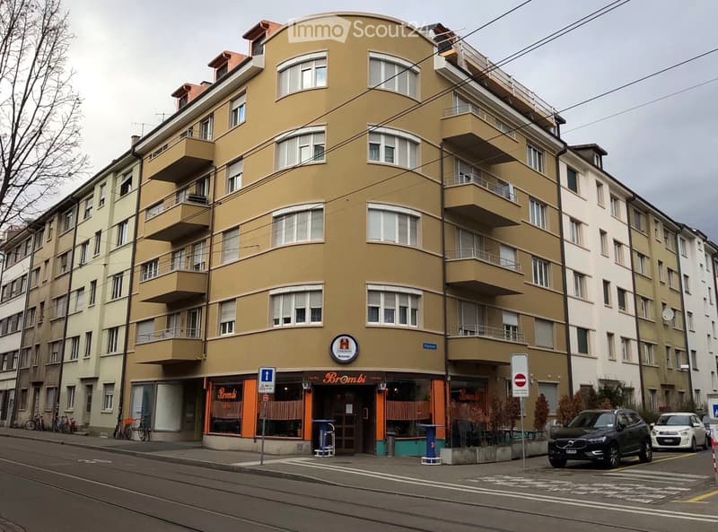 Multi-story residential building with balconies, storefront labeled "Brombi" at the ground level, and parked cars on the street.