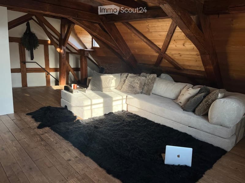 Cozy attic living space with exposed wooden beams, a large white sectional sofa, fur rugs, and a laptop on the floor.