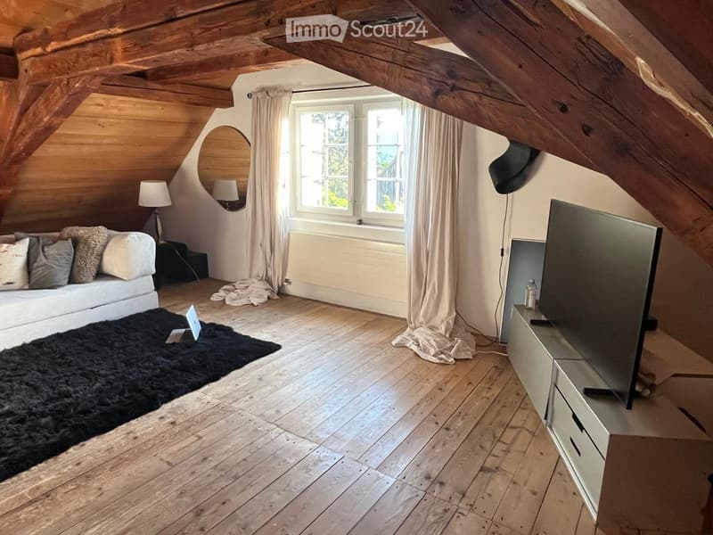 Cozy attic room with wooden beams, exposed wooden walls, large window, sofa, laptop, mirror, lamp, and television.