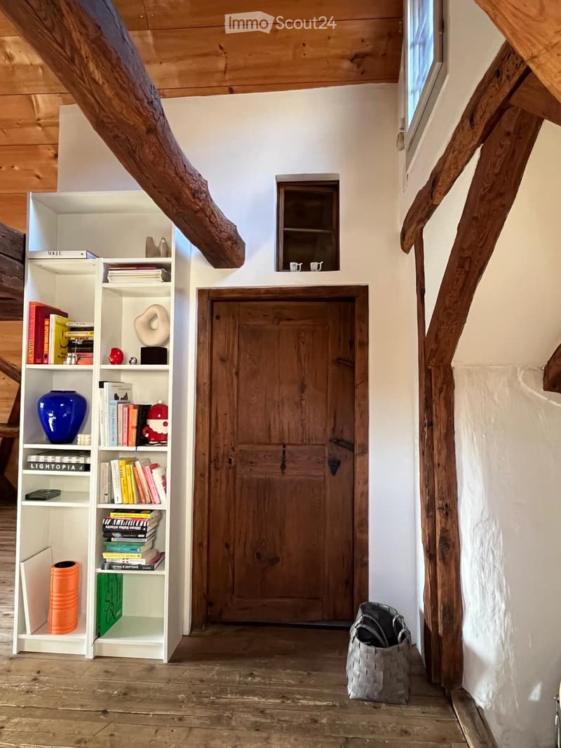 A rustic hallway with wooden beams, a white bookshelf filled with books and decorative items, a wooden door, and a woven basket on the floor.