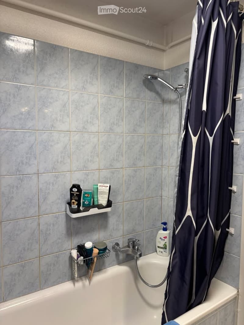 A modern bathroom with a bathtub, showerhead, and a dark blue shower curtain. The walls are tiled in light blue, and there are shelves holding various toiletries.