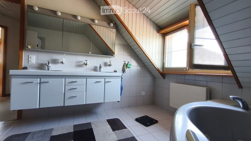 Modern bathroom with a double sink, large mirror, bathtub, and radiator. The room features white tiles, a window with blinds, and a patterned rug on the floor.
