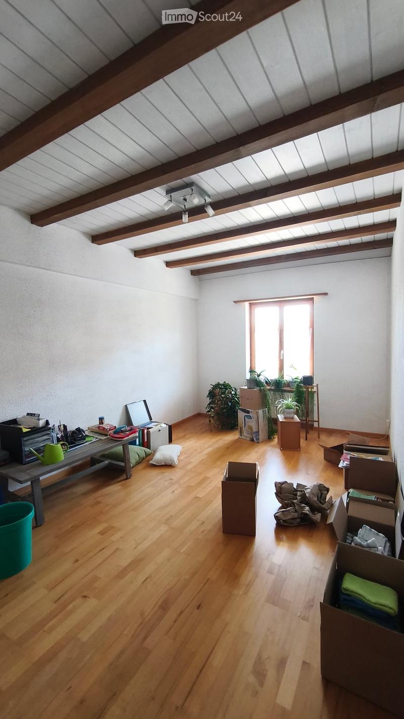 Empty room with wooden flooring, white walls, exposed wooden beams on the ceiling, and a large window letting in natural light. There are some boxes and items scattered around, indicating it is in the process of being unpacked or organized.
