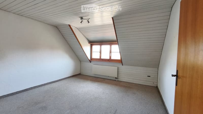 Empty attic room with sloped ceilings, white walls, wooden door frame, and a single window with wooden frame. The floor is carpeted, and there is a radiator under the window.