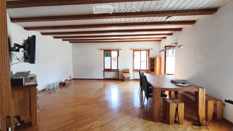Bright room with wooden floors, exposed wooden beams, large windows, a long wooden table with chairs, a laptop, a TV screen, and a small cabinet.