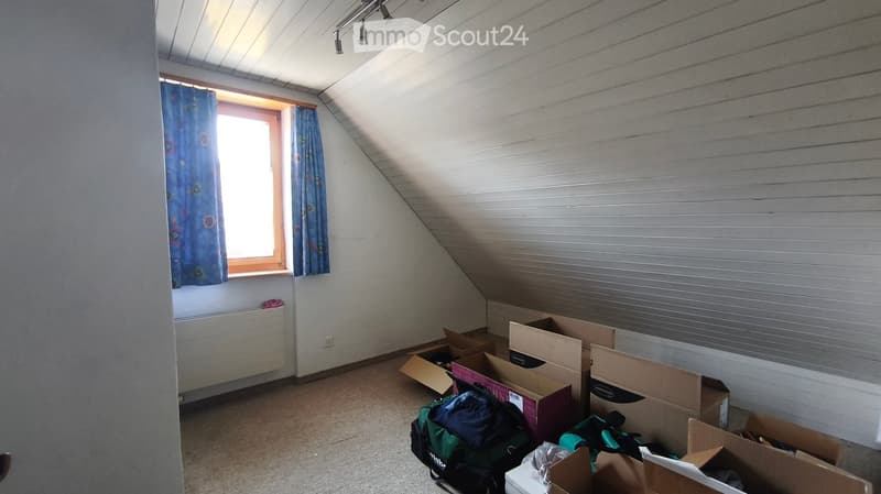 Empty attic room with slanted ceilings, exposed wooden beams, a window with blue curtains, and several cardboard boxes and bags on the floor.