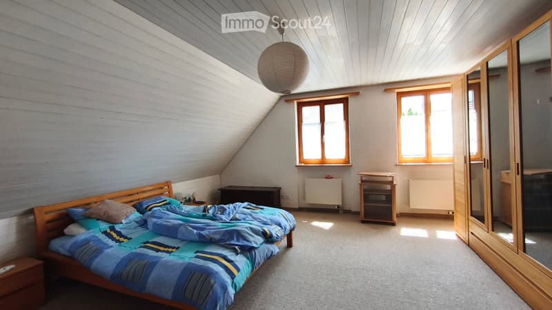 Bright attic bedroom with wooden slanted ceilings, large windows, a bed with blue and yellow bedding, a nightstand, a mirror wardrobe, and a small table.