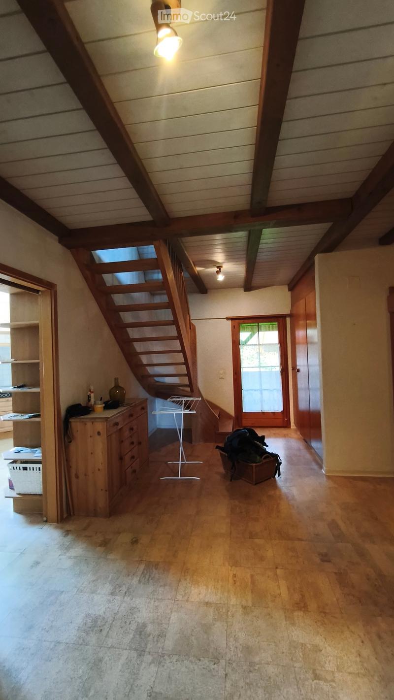 The image shows a spacious, well-lit hallway with wooden flooring and exposed wooden beams on the ceiling. A wooden staircase leads upwards, and a small wooden drawer unit is positioned near the staircase. A vase is placed on top of the drawer unit, and a