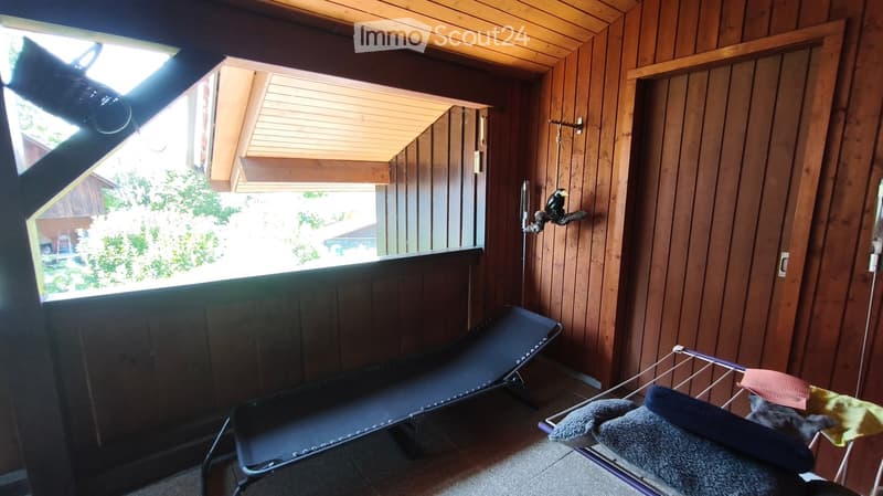 A small, wooden-paneled room with a large window offering a view of greenery. Contains a single bed and a laundry rack with folded clothes.