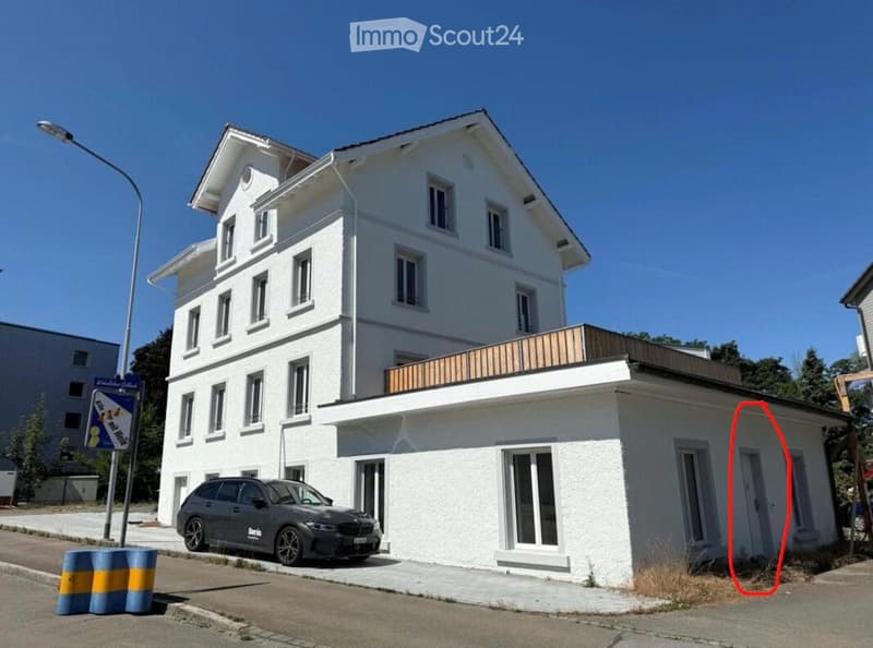 Two-story white building with multiple windows and a balcony on the roof. A car is parked in front.
