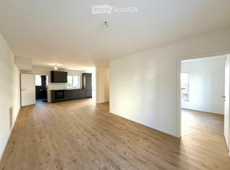 Bright, open-plan living and dining area with wooden flooring, white walls, and modern kitchen cabinets, sink, oven, and fridge visible in the background.