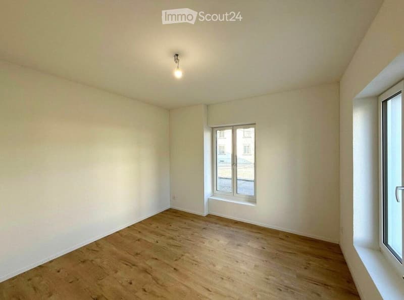 Empty room with wooden flooring, white walls, and two windows allowing natural light.