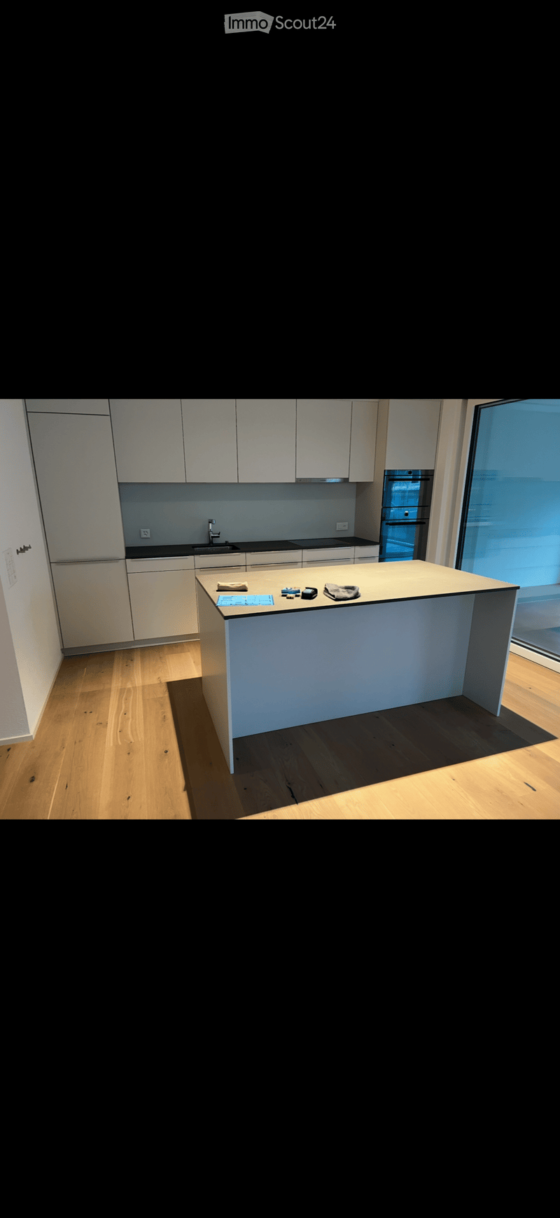 Modern kitchen with white cabinets, a central island, and a sink area. The floor is wooden, and the space is well-lit with natural light from a large window.