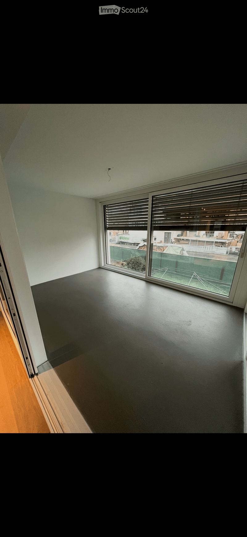 Empty room with dark grey walls and floor, large window with blinds, wooden flooring in adjacent area.