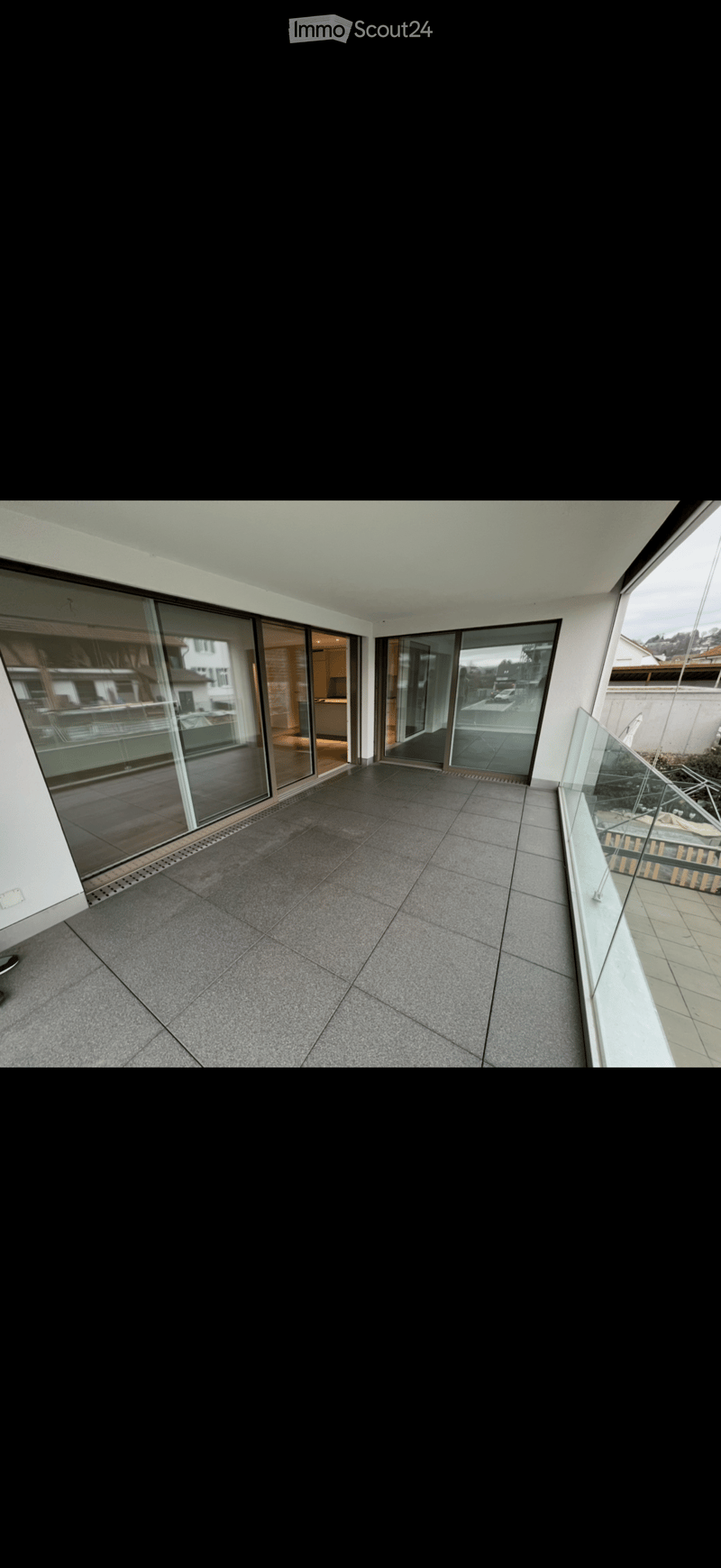 Modern balcony with grey tiled floor, large glass sliding doors, and a glass railing offering views of the surrounding area.