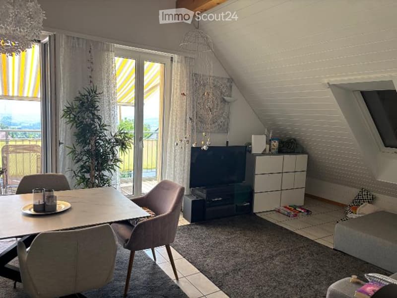 Bright living room with a modern aesthetic, featuring a grey sofa, a wooden dining table with chairs, a television, a small cabinet, and a potted plant near the large window with yellow blinds. The room has exposed wooden beams on the ceiling and a cozy r