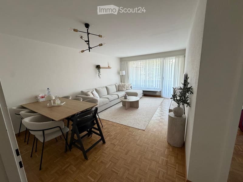 Modern living room with a white sofa, coffee table, dining table, chairs, and a plant in a vase. The room features wooden flooring and large windows with curtains.