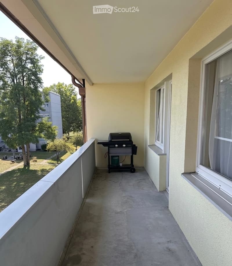 Balcony with a grill, concrete floor, beige walls, windows, and a view of a park with trees and a building.