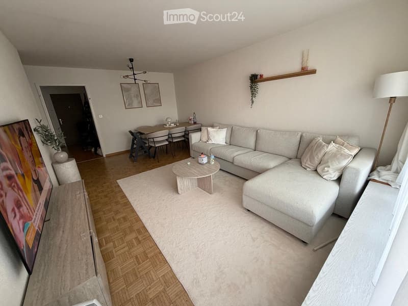 Modern living room with a white sectional sofa, coffee table, dining area with chairs, and a floor lamp. The space features wooden flooring and white walls.