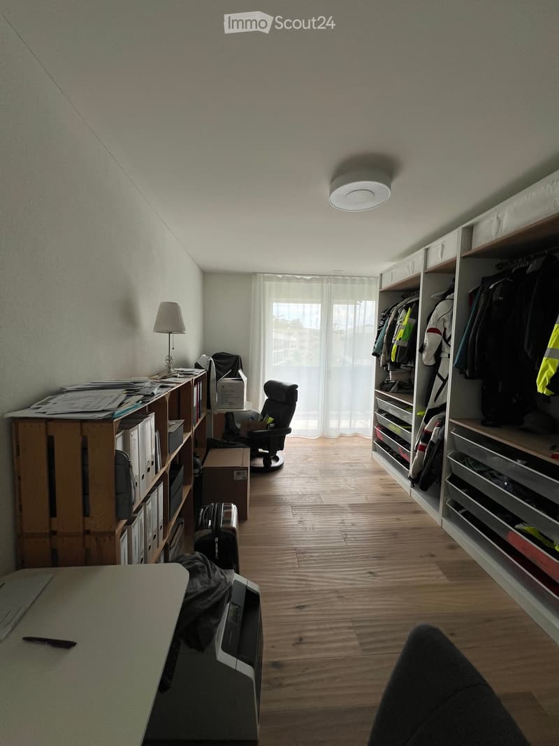 A furnished room with wooden flooring, a large window with white curtains, a chair, a table, a lamp, shelves, and a suitcase.