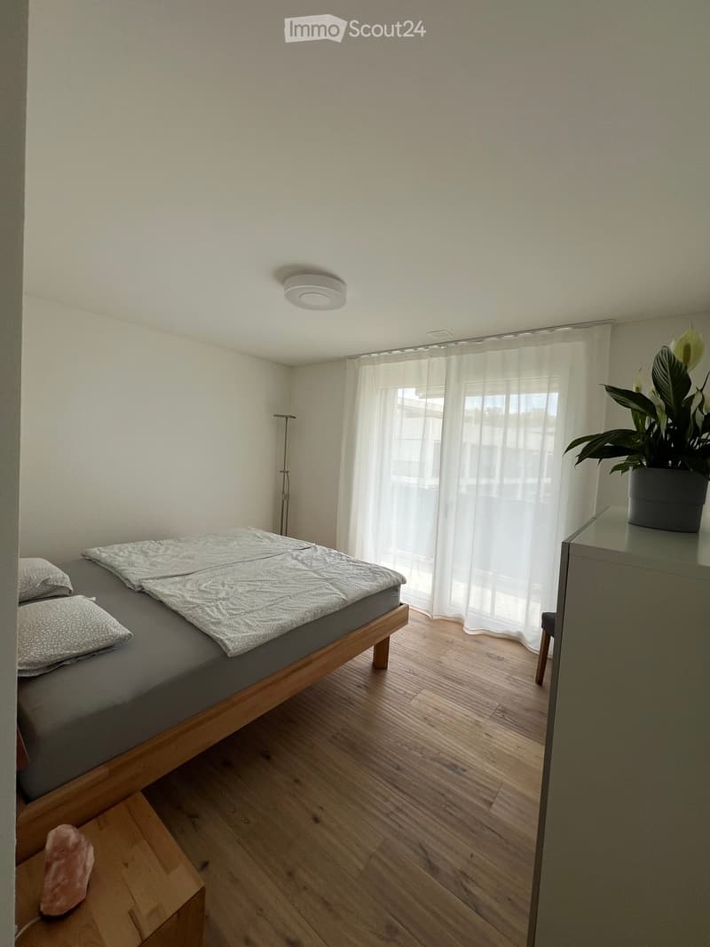 Minimalist bedroom with a wooden bed, grey bedding, white pillows, a nightstand with a plant, and a small stool with a decorative item.