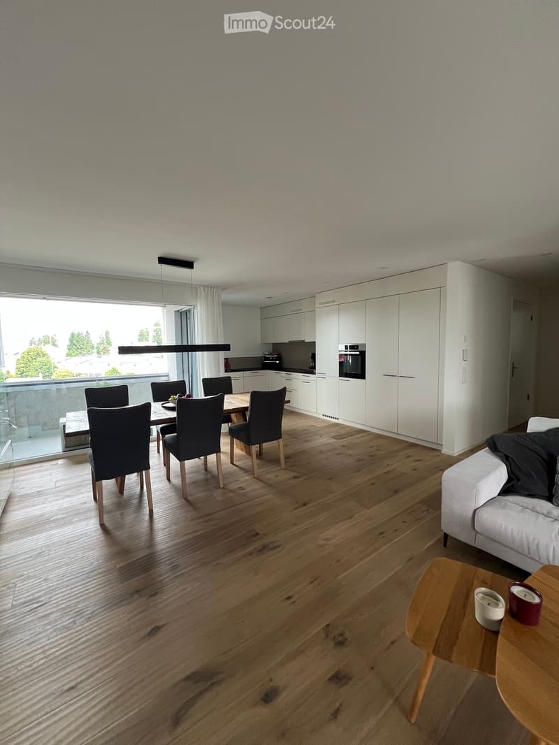 Open-plan living and dining area with wooden flooring, white cabinetry, a sofa, coffee table, dining table with chairs, and an oven in the background.