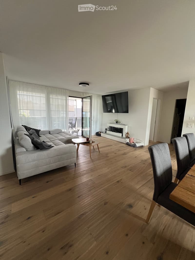 Modern living room with hardwood floors, large windows, a grey sectional sofa, a coffee table, a fireplace, and a TV mounted on the wall. A dining area with chairs and a table is visible.
