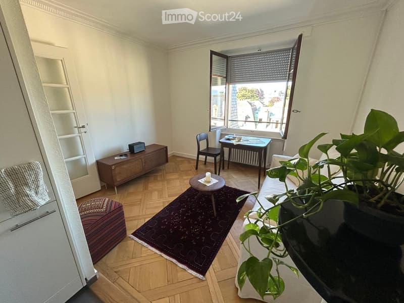 A modern living room with wooden flooring, a large window with blinds, a sofa, coffee table, small table, chair, plant, and a cabinet.