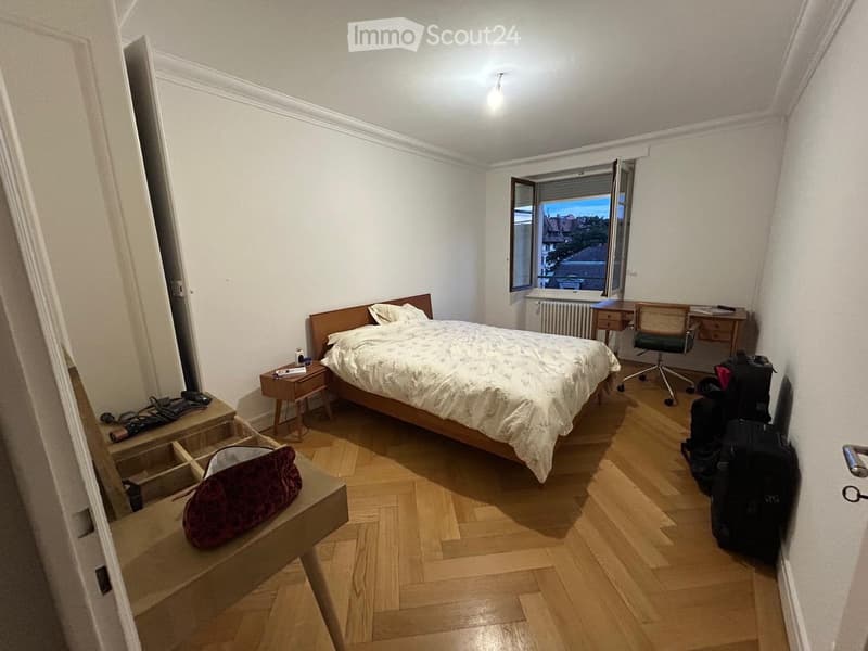 Cozy bedroom with wooden flooring, a double bed with white bedding, a nightstand, a desk with a chair, and a window offering a view of the city.