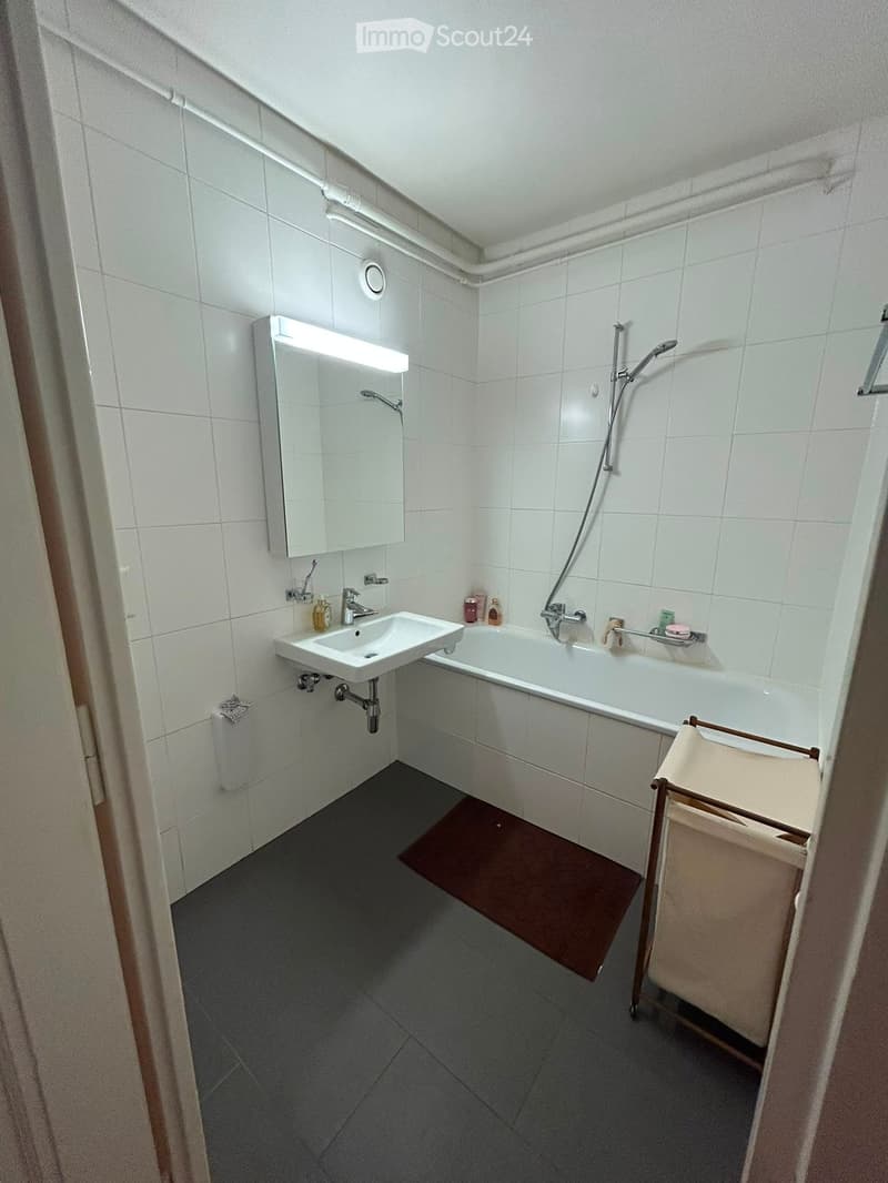 Modern bathroom with white tiles, a sink, bathtub, mirror, and a towel rack. Includes a bath mat on the floor.