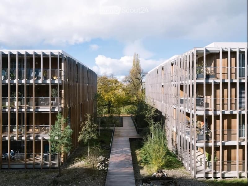 Modern multi-story residential building with wooden balconies and outdoor seating areas, surrounded by greenery and a wooden walkway.